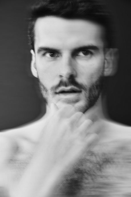 A black and white blurred photo of a shirtless man with a beard and mustache, looking directly at the camera with one hand near his chin.