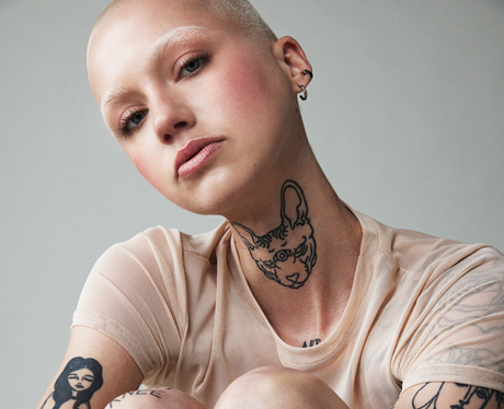 A joyful bald woman with pale skin laughing and touching her face, showcasing multiple tattoos on her arms and a prominent tattoo of a cat on her neck, wearing a light beige t-shirt against a light grey background.
