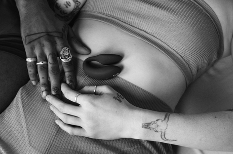  A black and white photo of two hands on a clothed torso, one with rings and a dinosaur tattoo, the other with a plus sign tattoo.